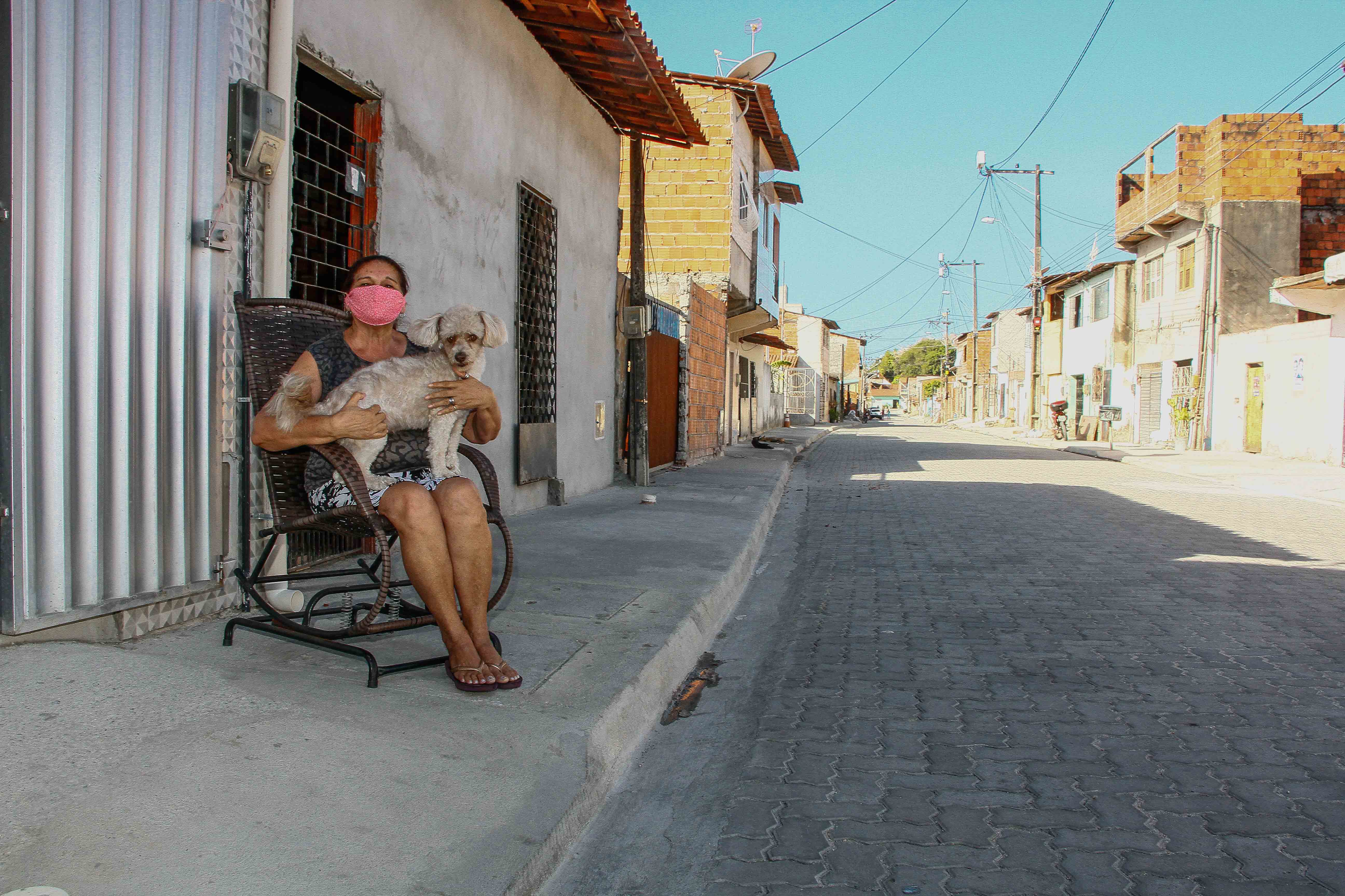 senhora de máscara sentada em cadeira sobre calçada com cachorro no colo e rua em frente rua com piso intertravado